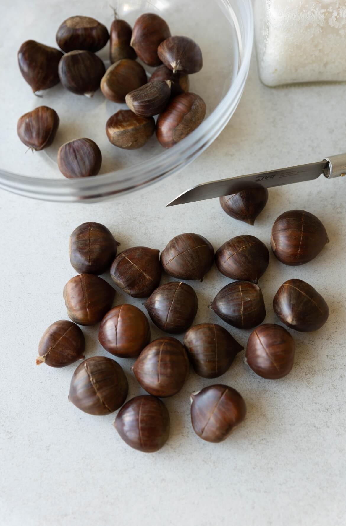 processo de preparação de castanhas, com várias castanhas dispostas sobre uma superfície clara. Algumas castanhas têm cortes feitos para facilitar o cozimento