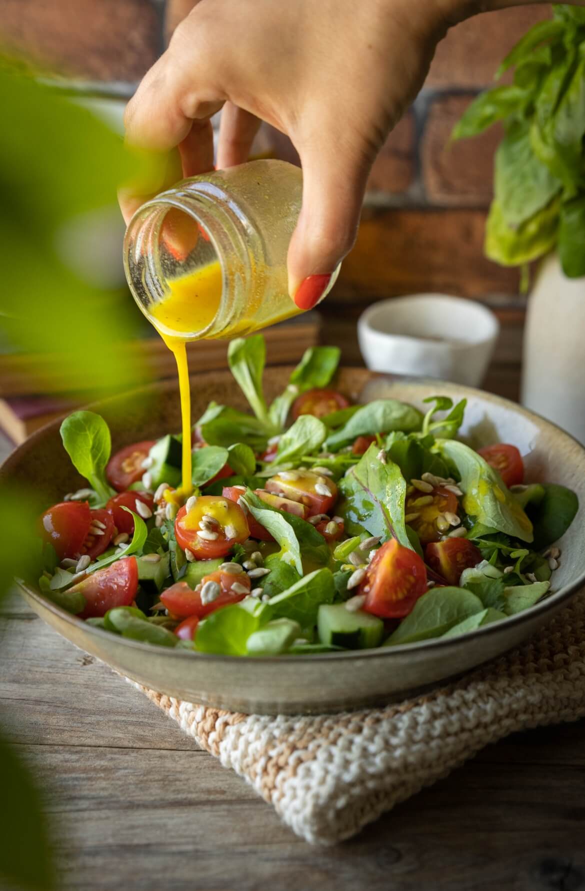 Salada de folhas verdes com canónigos, tomate cherry, pepino e sementes de girassol. Uma mão a verter vinagrete de mostarda e cúrcuma. 
