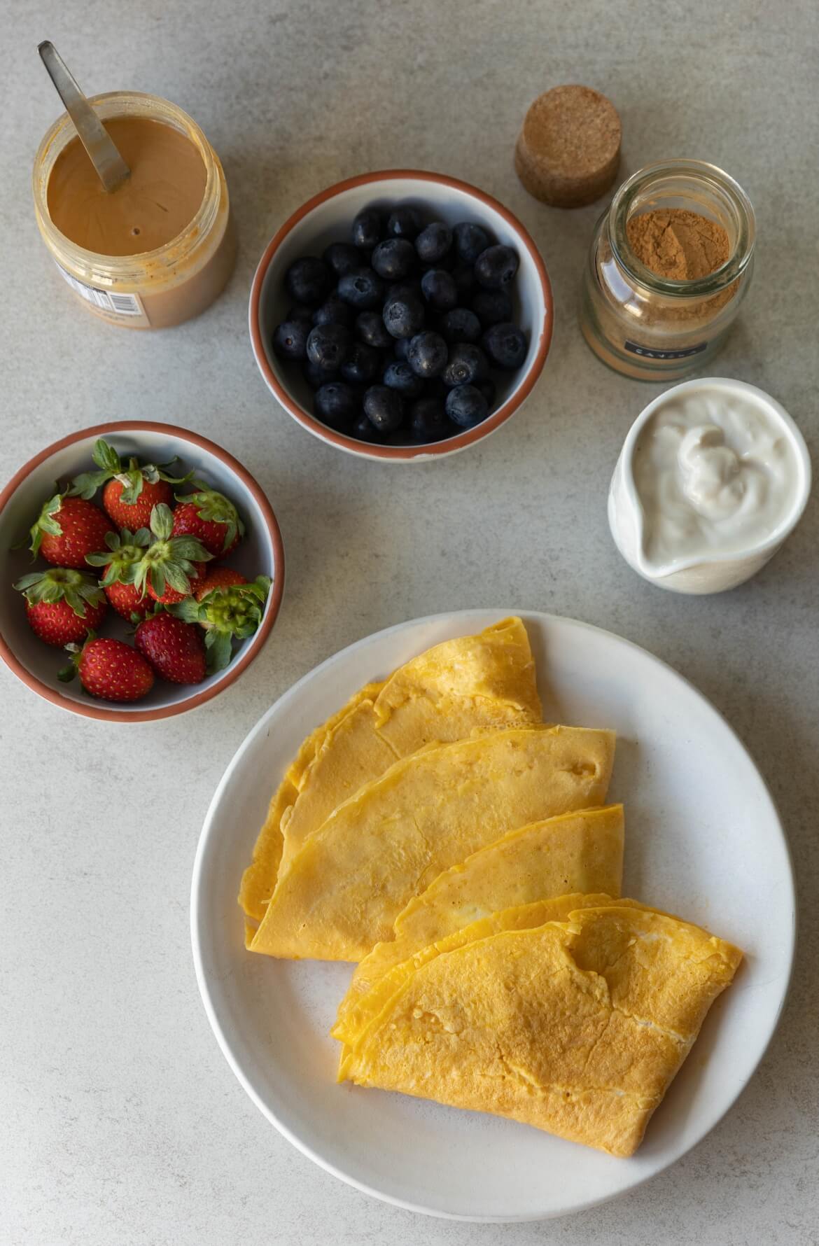 Ingredientes para crepioca doce: morangos, mirtilos, iogurte vegetal, canela e creme de amendoim, com crepiocas prontas no prato.