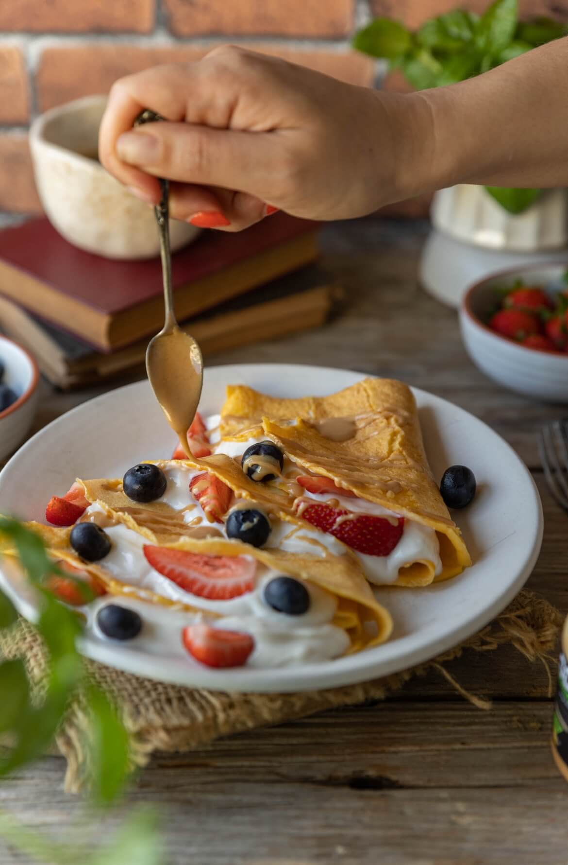 Mão a servir creme de amêndoa sobre crepioca doce com iogurte e frutos vermelhos.